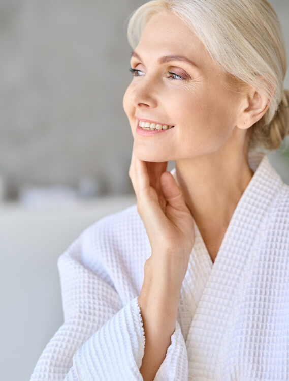 Headshot vertical portrait of smiling middle aged woman wearing bathrobe touching face at spa salon hotel looking away. Bodycare spa procedures skin care antiaging products concept.